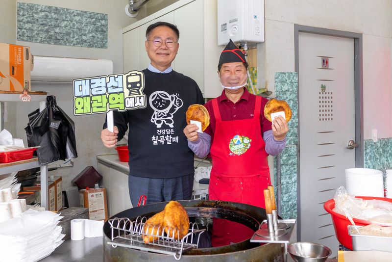 사진1김재욱 칠곡군수가 ‘대경선 왜관역에서’라는 문구가 적힌 팻말을 들고 웰빙왕호떡을 홍보하고 있다. 대경선을 타고 왜관역에 내려 호떡을 즐길 수 있다는 의미를 담은 촬영 장면.