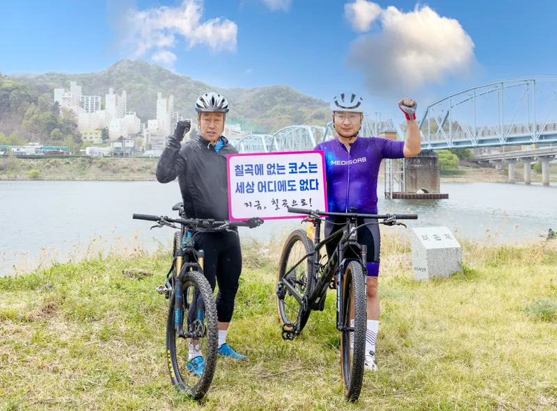 김태경 칠곡군 사이클연맹 회장(왼쪽)과 김원일 원바이오젠 대표가 6·25전쟁 당시 북한군의 진격을 막기 위해 폭파했던‘호국의다리’아래에서 기념촬영을 하고 있다. 두 사람은‘칠곡에 없는 코스는 세상 어디에도 없다’는 문구가 적힌 피켓을 들고, MTB 성지로 떠오른 칠곡의 자전거 코스를 알리고 있다.